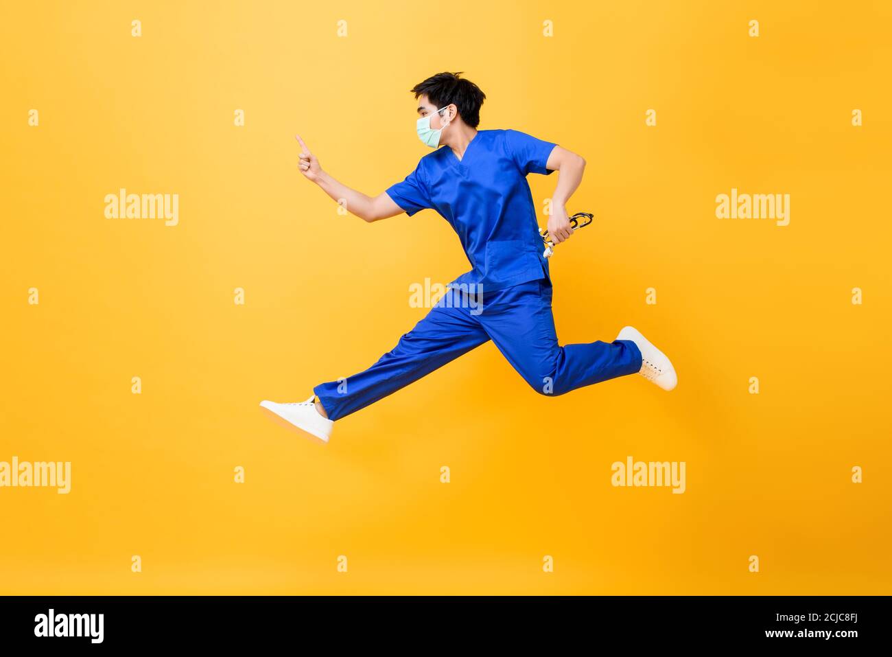 Young Asian male doctor wearing medical mask jumping and pointing hand up isolated in yellow studio background Stock Photo