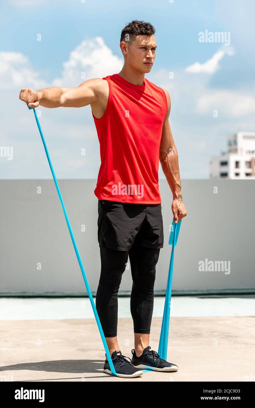 Handsome sports man doing shoulder lateral raise exercise with resistance band outdoors on rooftop in the sun Stock Photo