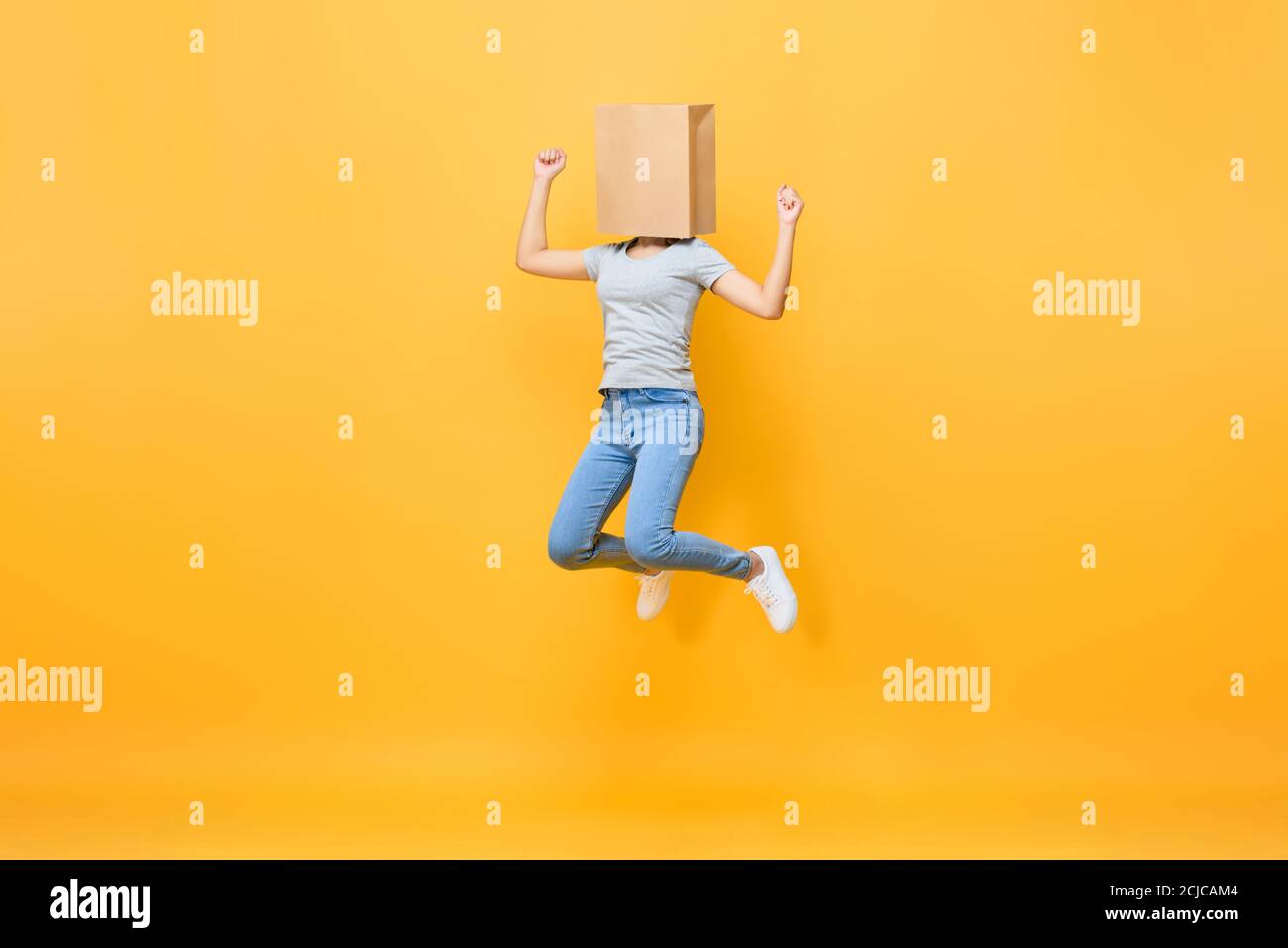Fun portrait of faceless anonymous woman covering head with paper bag jumping in mid-air isolated on yellow studio background Stock Photo