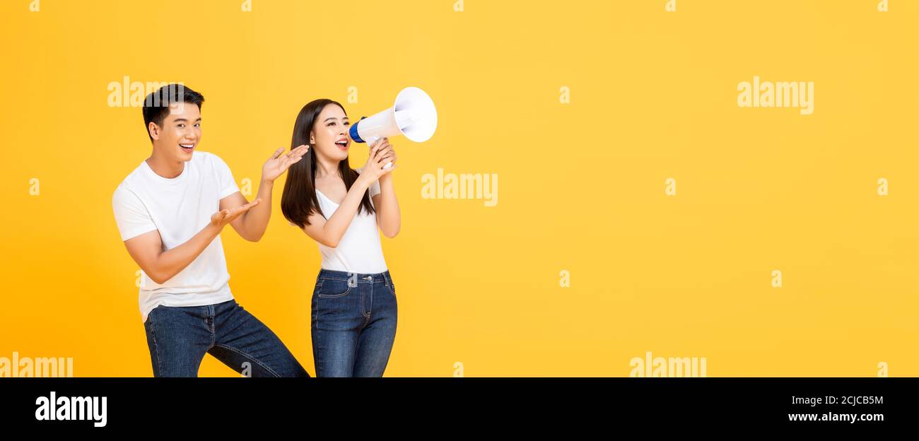 Cheerful portrait of smiling happy young Asian couple making announcement and presenting in isolated studio banner yellow background with copy space Stock Photo