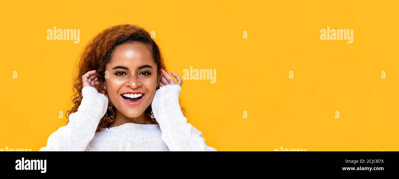 Close up portrait of cheerful young African American woman smiling while touching her ears in isolated studio yellow banner background with copy space Stock Photo