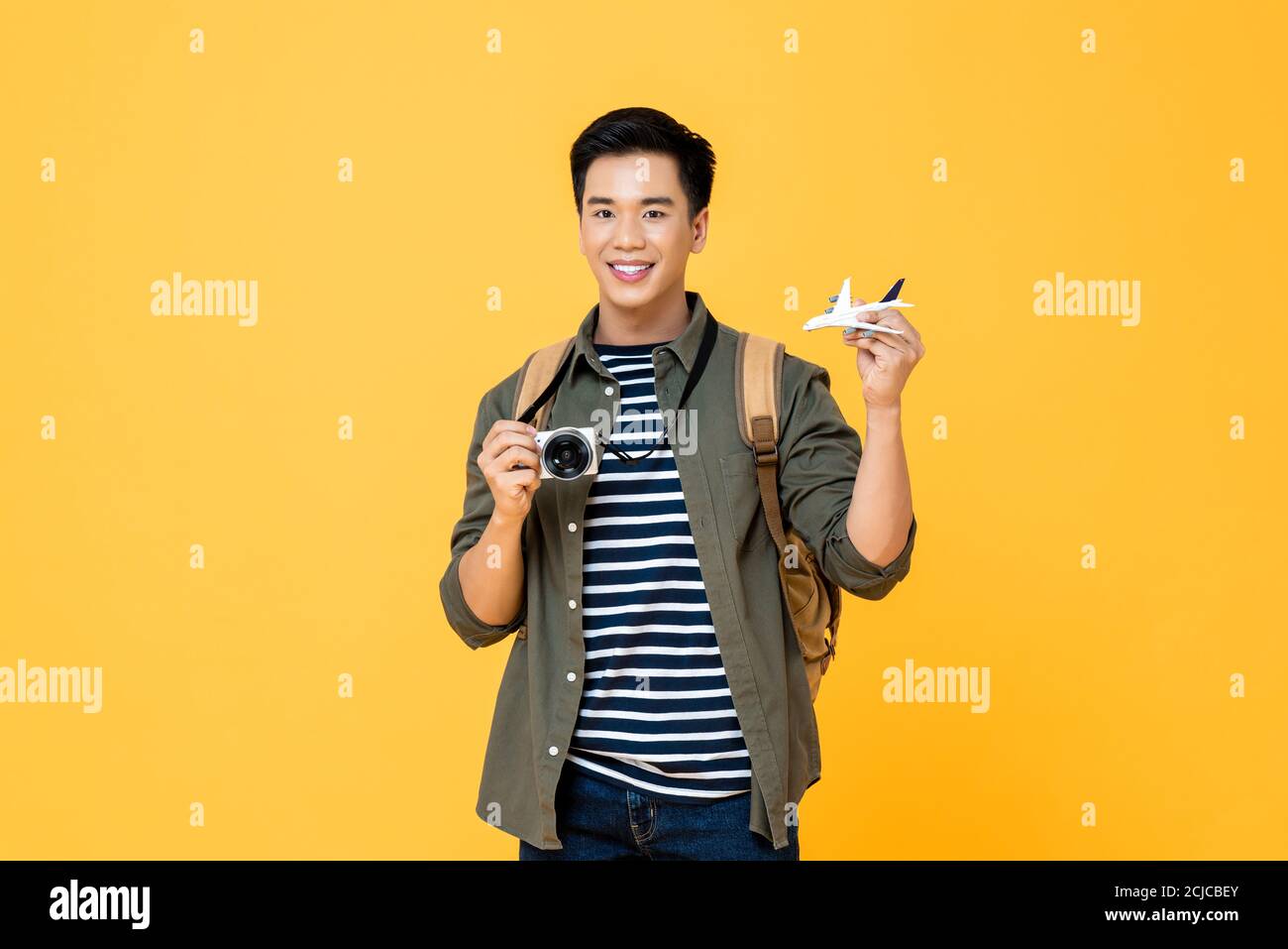 Portrait of young handsome smiling Asian male tourist backpacker holding plane model and camera isolated on studio yellow background Stock Photo