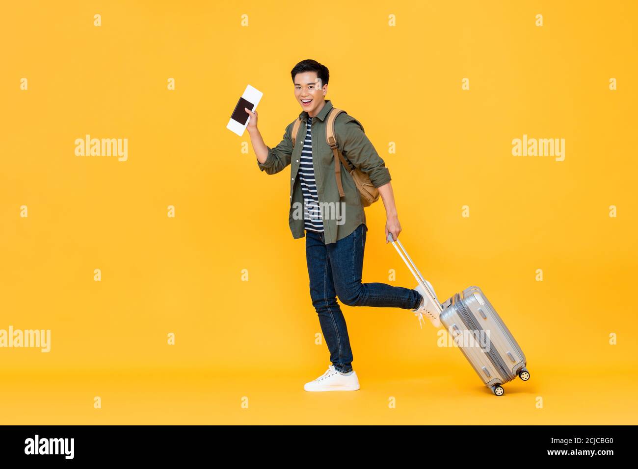 Full length portrait of smiling happy handsome young Asian man tourist with passport and luggage ready to travel on vacations isolated in yellow studi Stock Photo