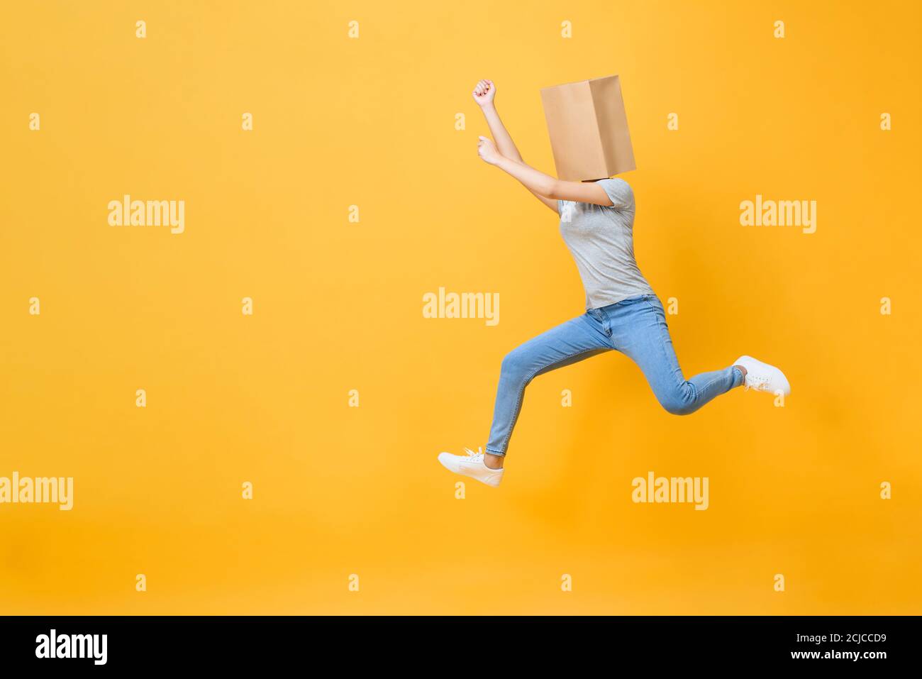Fun portrait of anonymous woman with head covered with paper bag jumping in mid-air isolated on yellow studio background Stock Photo