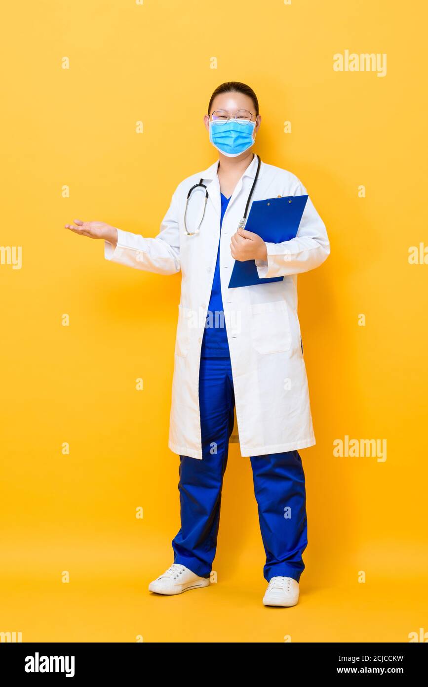 Full length portrait of young Asian woman doctor wearing face mask with open palm gesture in isolated studio yellow background Stock Photo