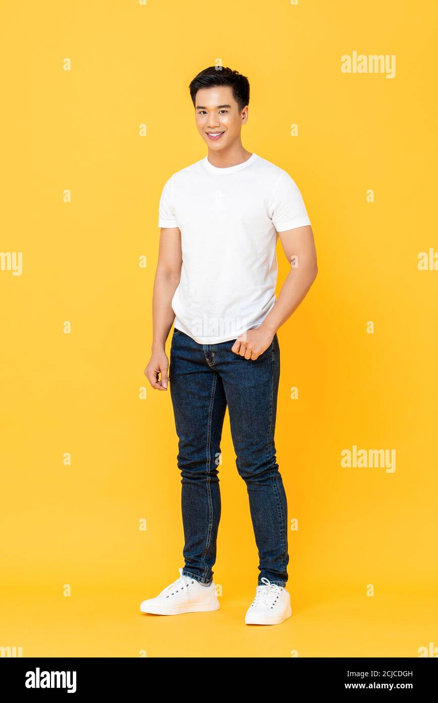Full length portrait of smiling young handsome Asian man standing with one hand in pocket isolated on yellow studio background Stock Photo