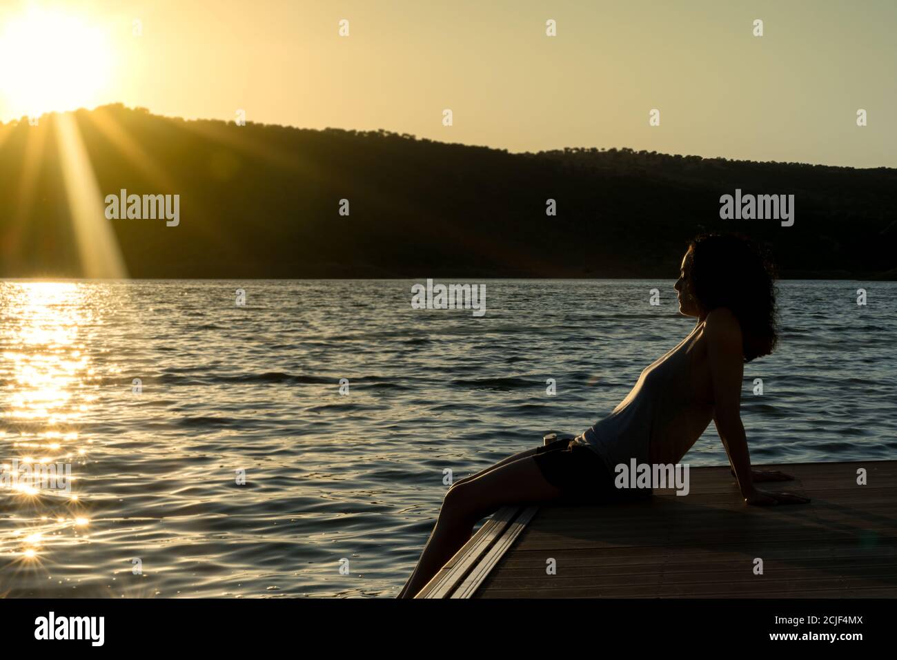 young girl with curly hair sitting on a pier looking at the horizon in a sunset Stock Photo