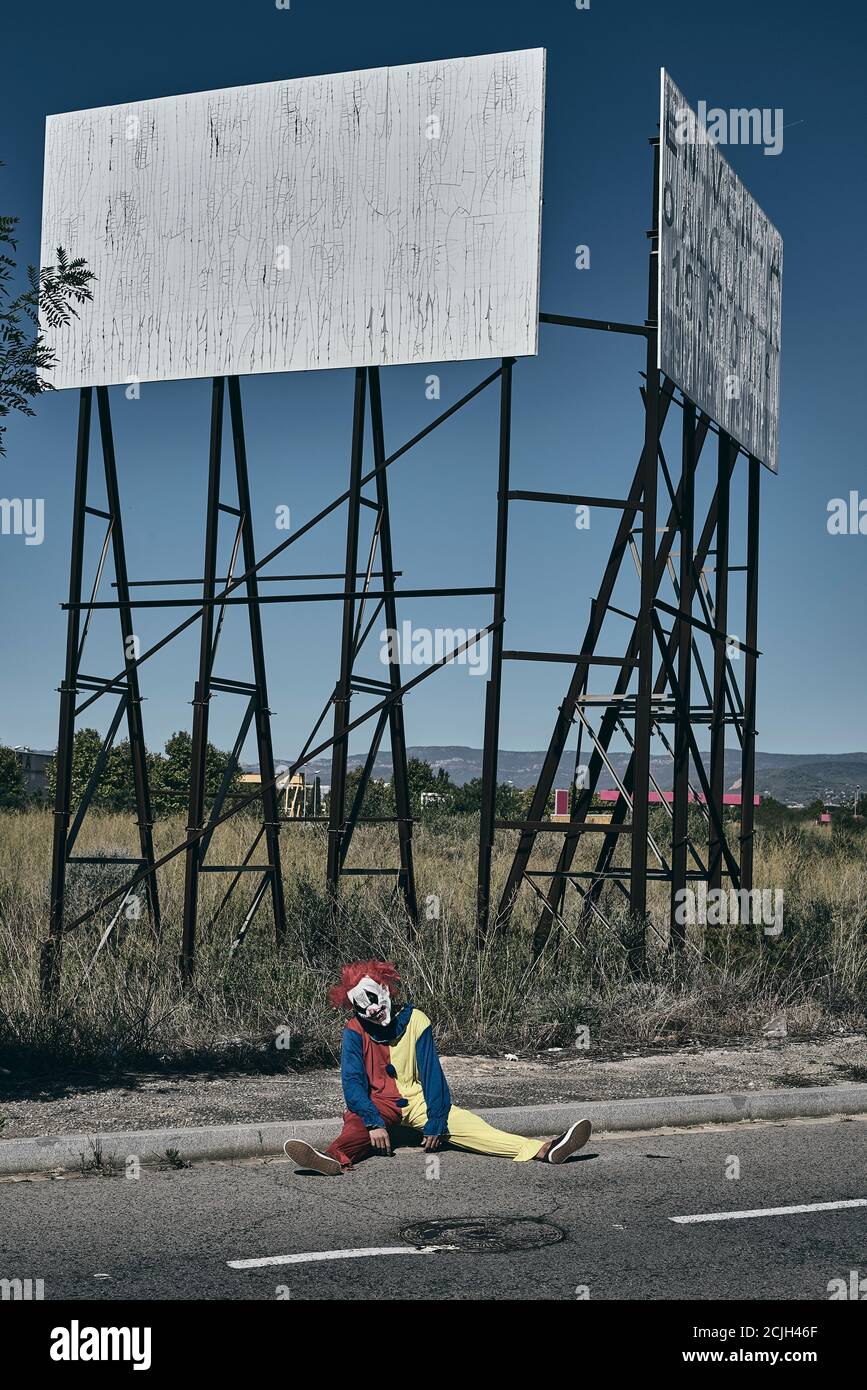 a scary evil clown, wearing a yellow, red and blue costume outdoors, sitting on the ground on a rural road in front of the rusty structure of an aband Stock Photo