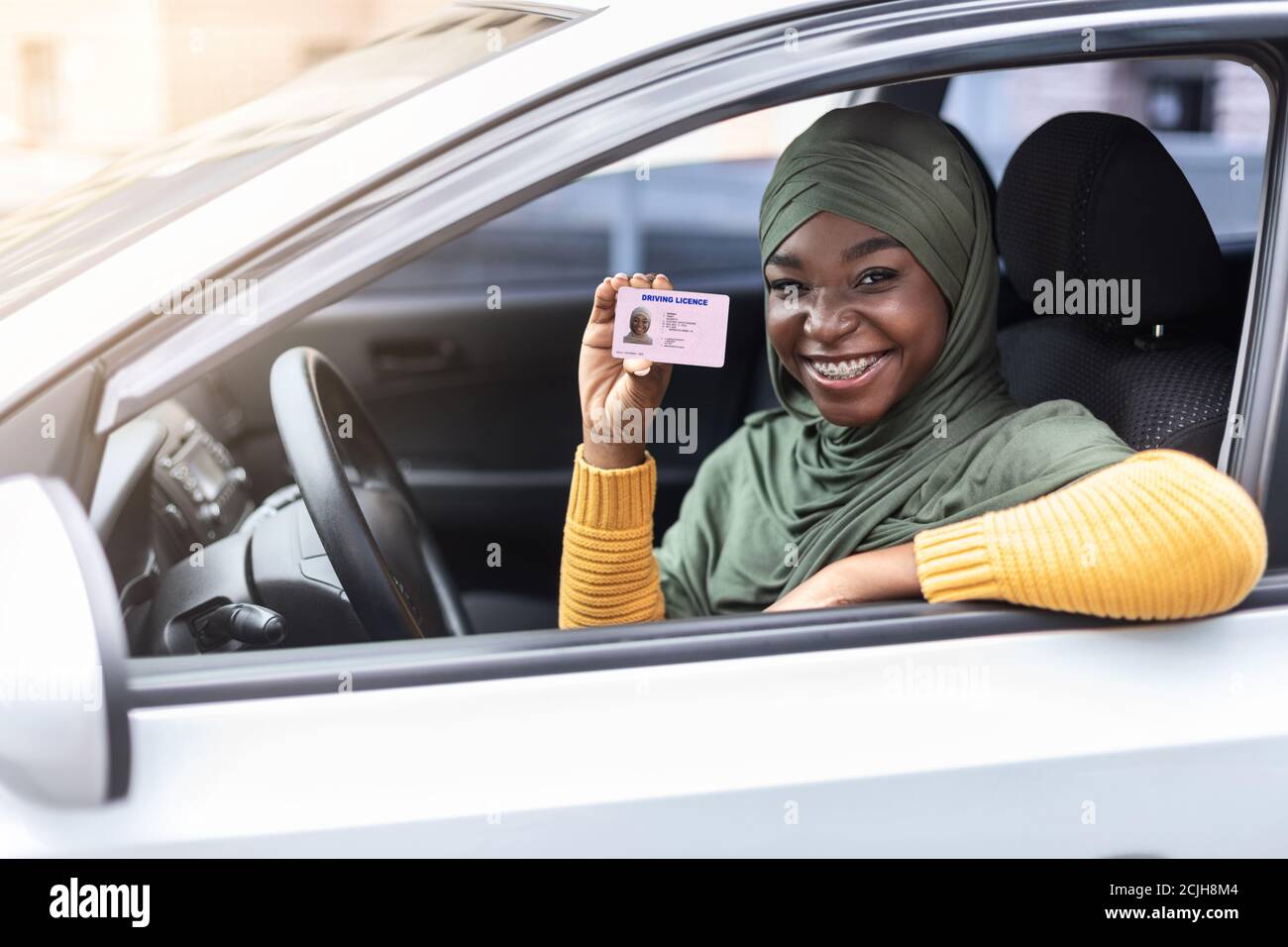 Happy black muslim woman in hijab sitting in car, showing driver license Stock Photo