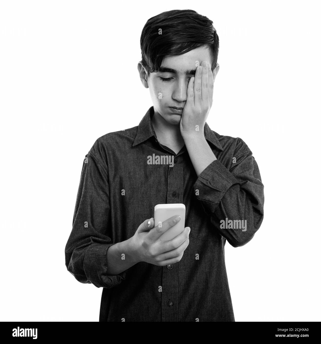 Studio shot of young stressed Persian teenage boy using mobile phone while covering face Stock Photo