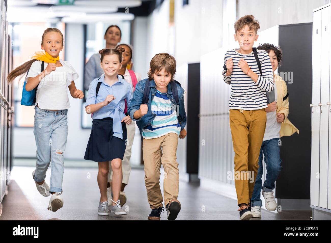 excited multicultural pupils running along school corridor with teacher on background Stock Photo