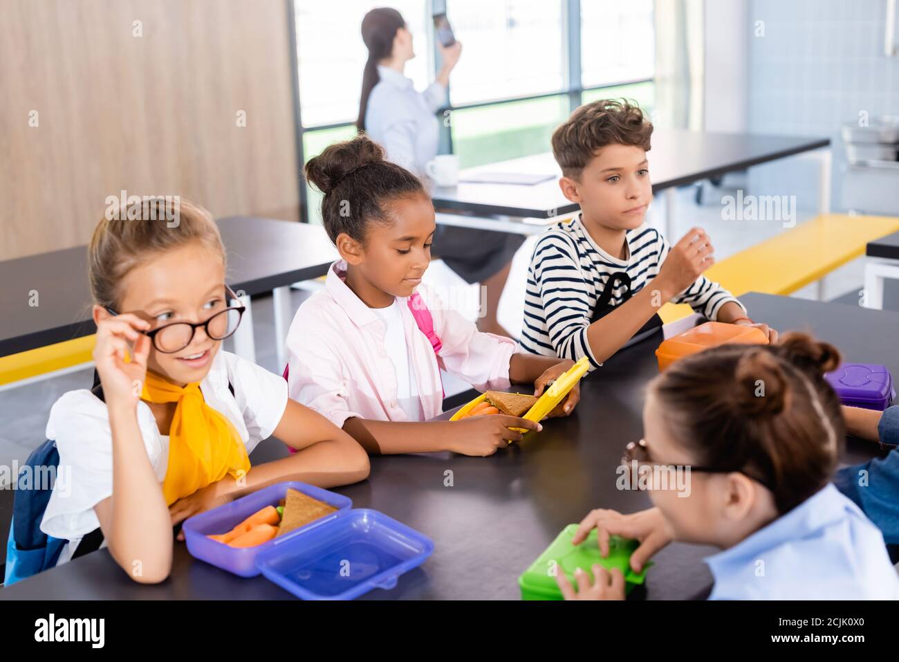 schoolgirls talking while sitting in school eatery with multicultural classmates and teacher on background Stock Photo