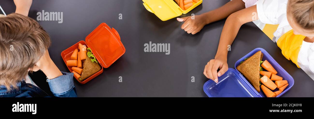 overhead view of multicultural pupils near lunch boxes with sandwiches and fresh carrots, horizontal image Stock Photo