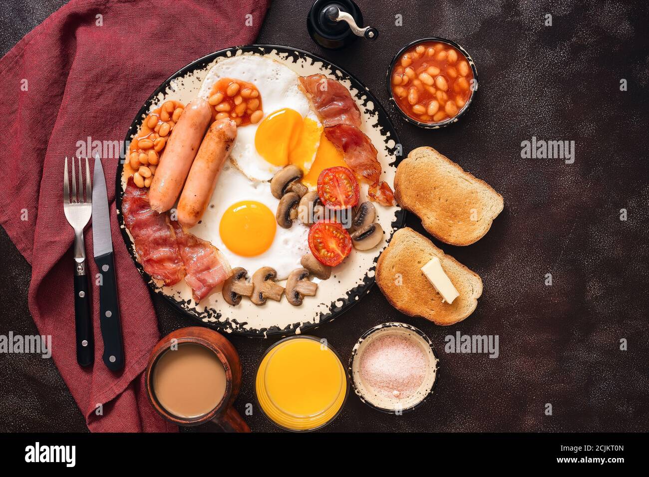 English traditional breakfast on a dark brown rustic background. Top view, flat lay. Fried eggs with bacon, sausages and beans, coffee, orange juice. Stock Photo