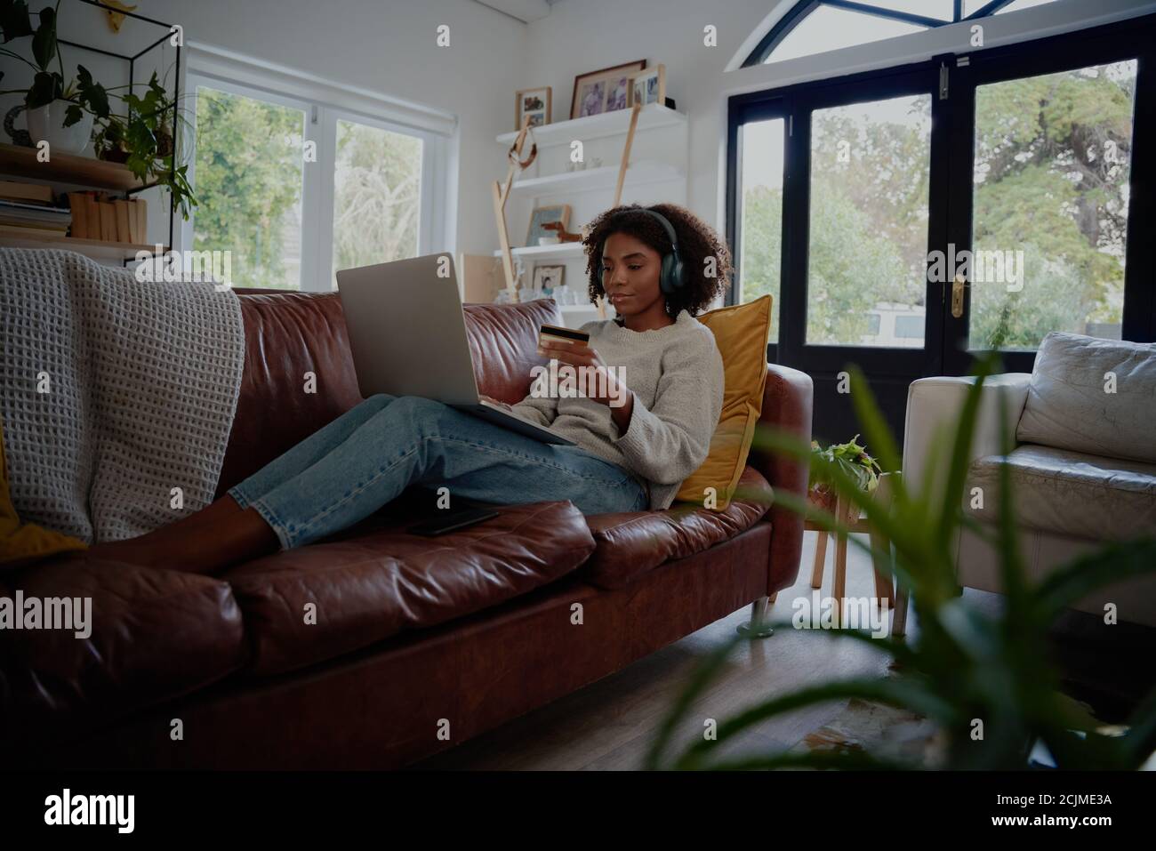 African American young woman relaxing on couch listening to music while making online payment using bank card and laptop Stock Photo