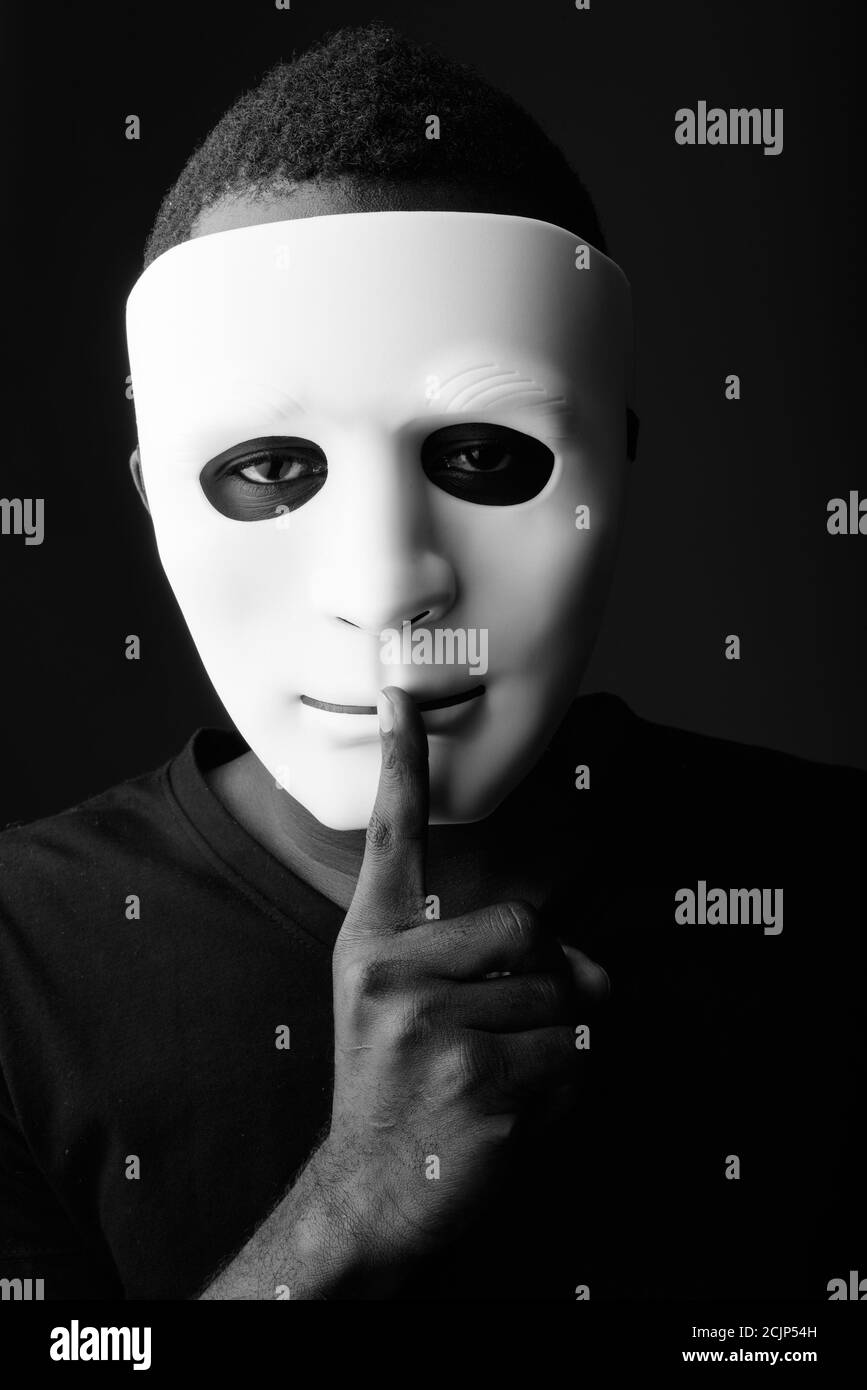 Studio shot of young black African man in dark room wearing mask and holding finger on lips Stock Photo