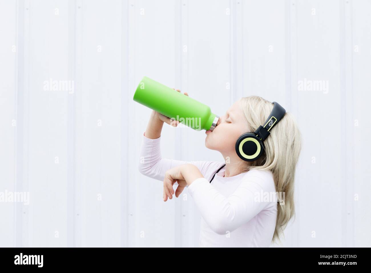 Young blond girl on white background drink fresh spring water from green reusable bottle. Healthy lifestyle in summer city. Stock Photo