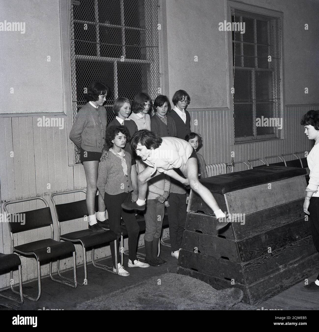 1960s, historical, a girl leaping over a traditional wooden vaulting horse or box inside a youth club, Bowhill, Fife, Scotland, watched by other girls standing on chairs. The girls club movement in Scotland has long roots having started as far back as the 1860s. Youth clubs play an important role in communities giving youngsters a place to play, meet and take part in sporting activities. Stock Photo