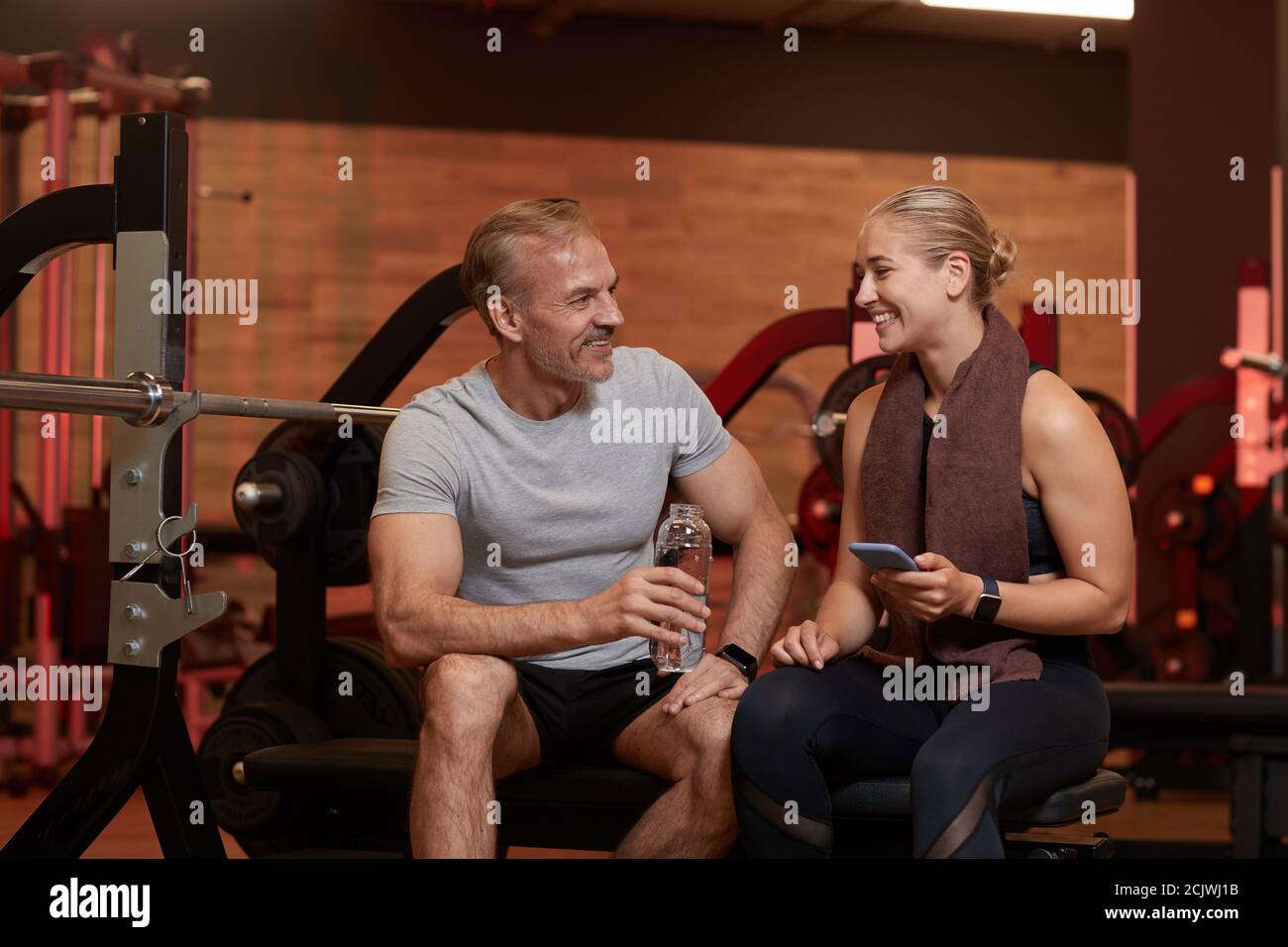Happy couple drinking water and using mobile phone and talking to each other after sports training in gym Stock Photo