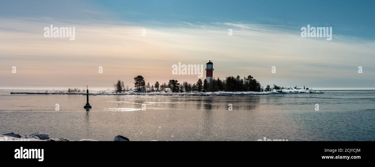 Panorama of red white lighthouse in middle of frozen, covered with snow island at cold Baltic Sea, partly open water, thin ice reflecting day light. B Stock Photo