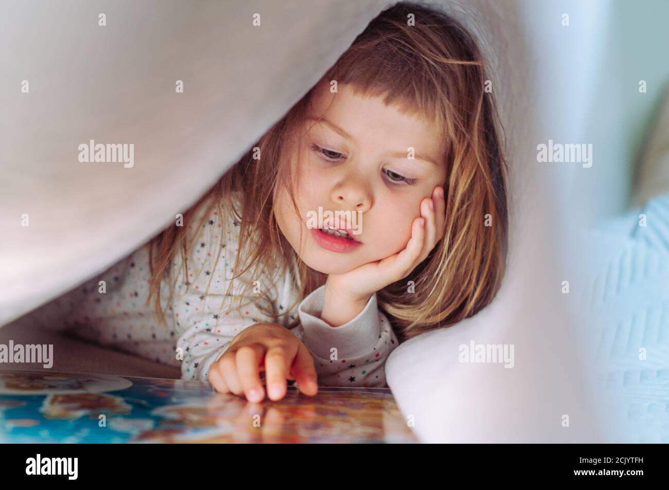Beautiful little girl reading book lying on the bed under the blanket