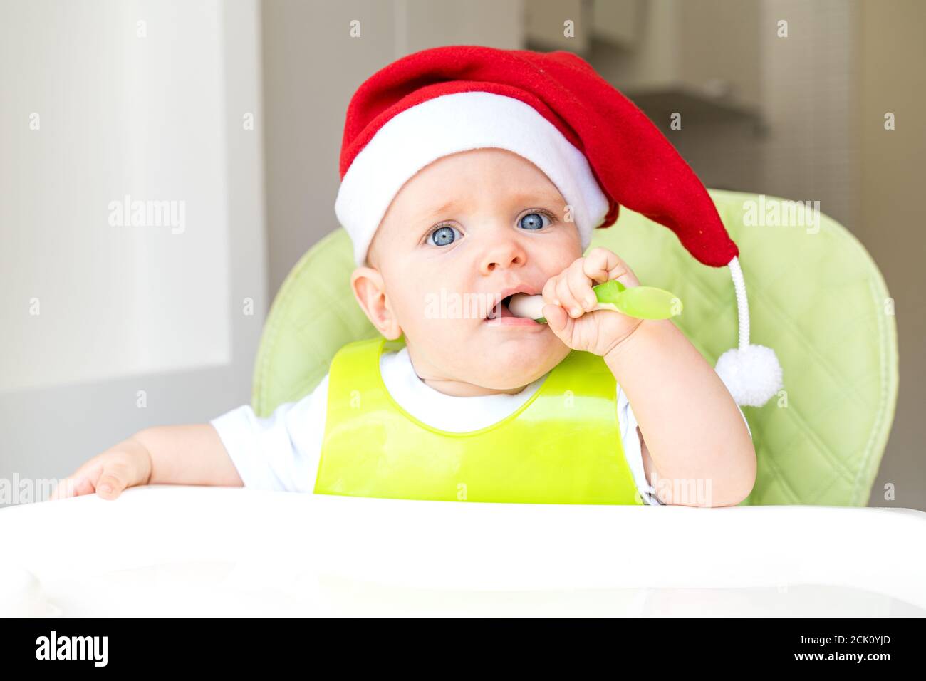 a baby in a Santa Claus hat is sitting in a high chair and eating with a spoon Stock Photo