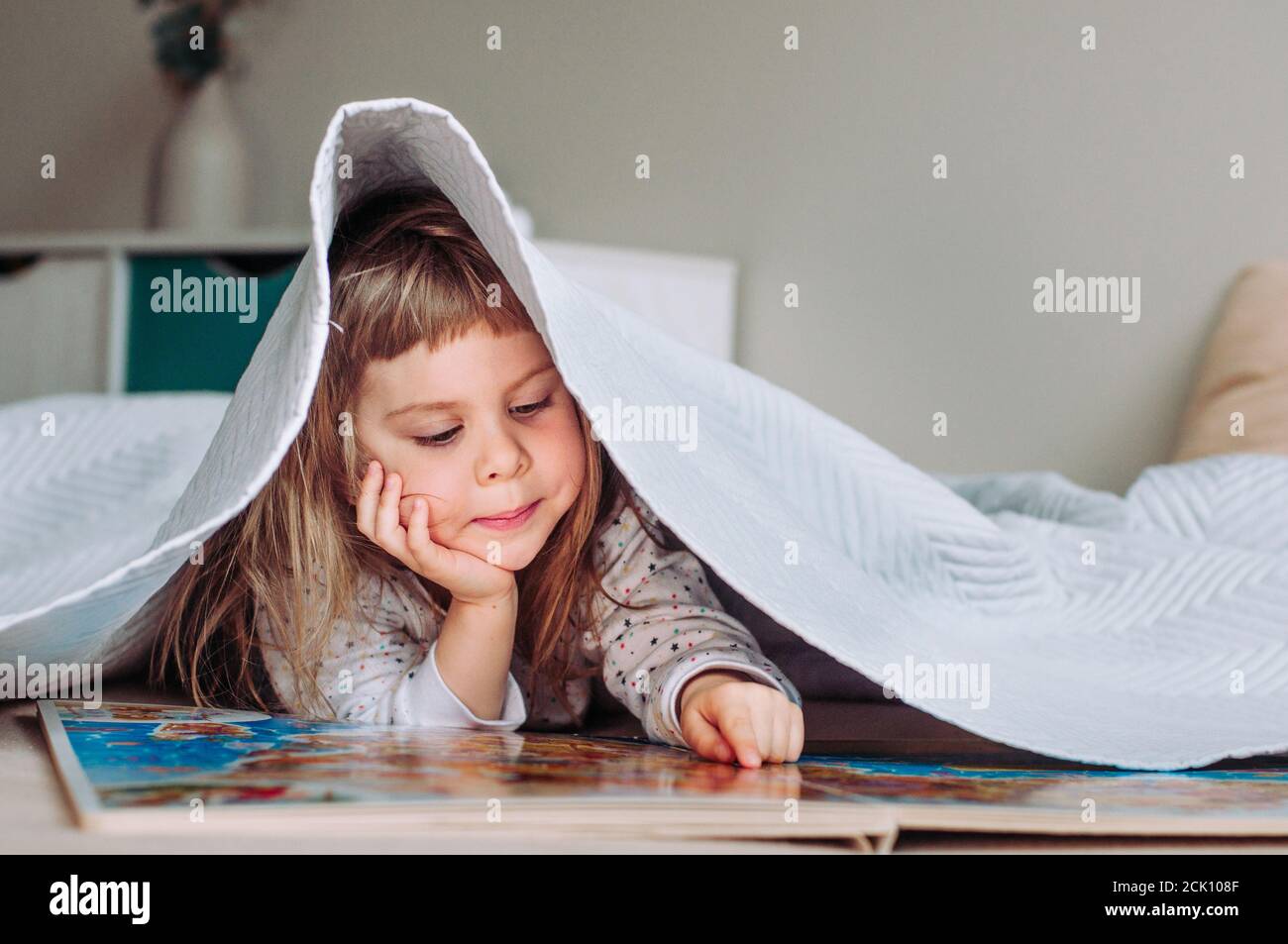 Beautiful little girl reading book lying on the bed under the blanket