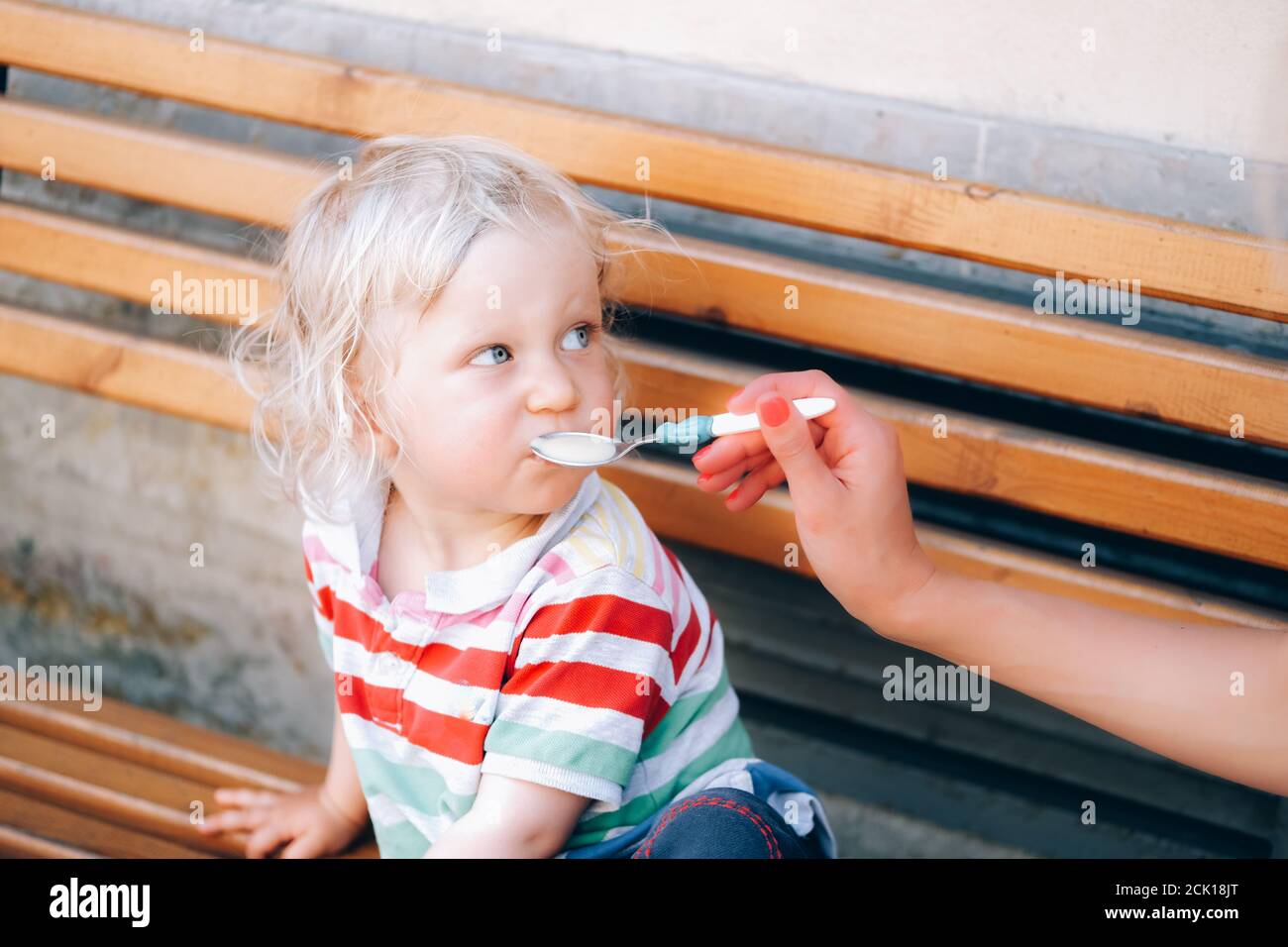 Cute little blond boy with blue eyes is eating outside from the spoon, held by his mother. Feeding a baby during the city walk on the banch. Stock Photo