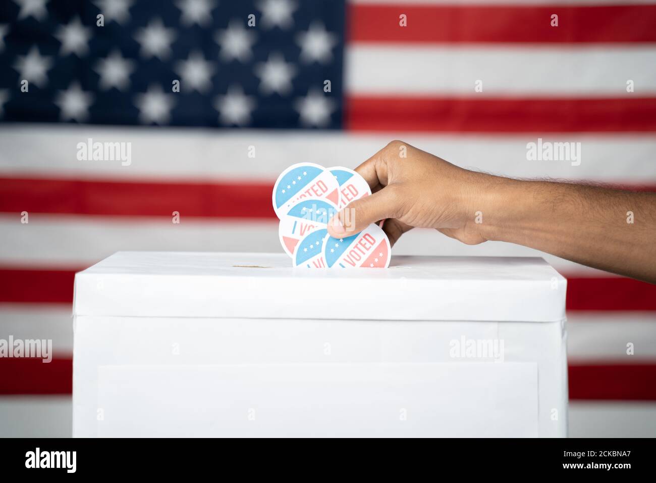 Close up of Hands dropping multiple I Voted sticker inside Ballot box with US flag as background, Concept of fraud in USA elections. Stock Photo