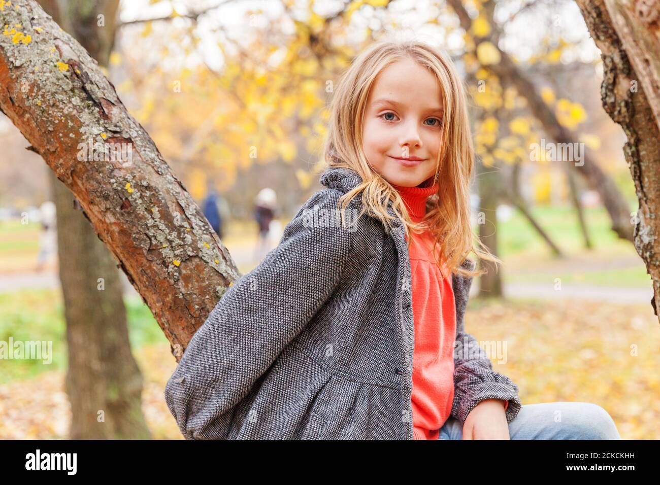 Happy young girl smiling and sitting on tree in beautiful autumn park on nature walks outdoors. Little child playing in autumn fall orange yellow background. Hello autumn concept Stock Photo