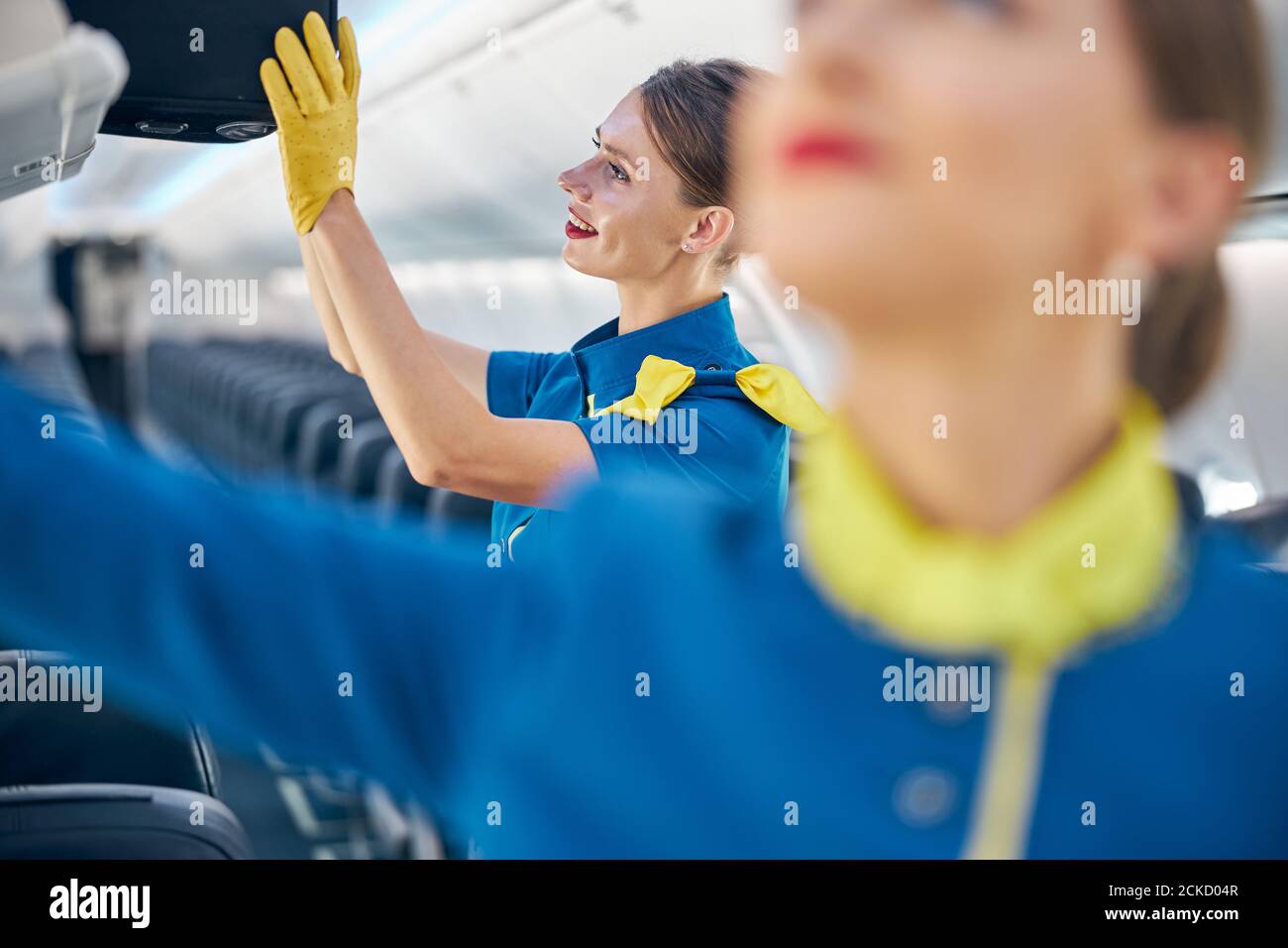 Close up portrait of smiling cheerful female wearing flight attendant blue uniform holding in hand black rolling bag while putting it on the cabin com Stock Photo