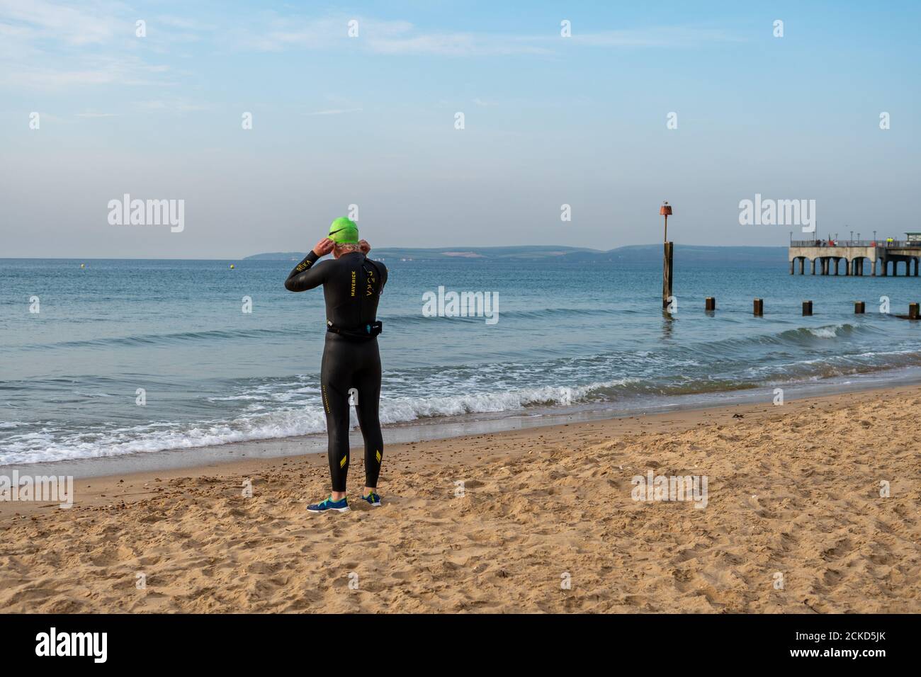 A sea swimmer wearing a wetsuit pulling on a green swimming cap while preparing to enter the sea Stock Photo