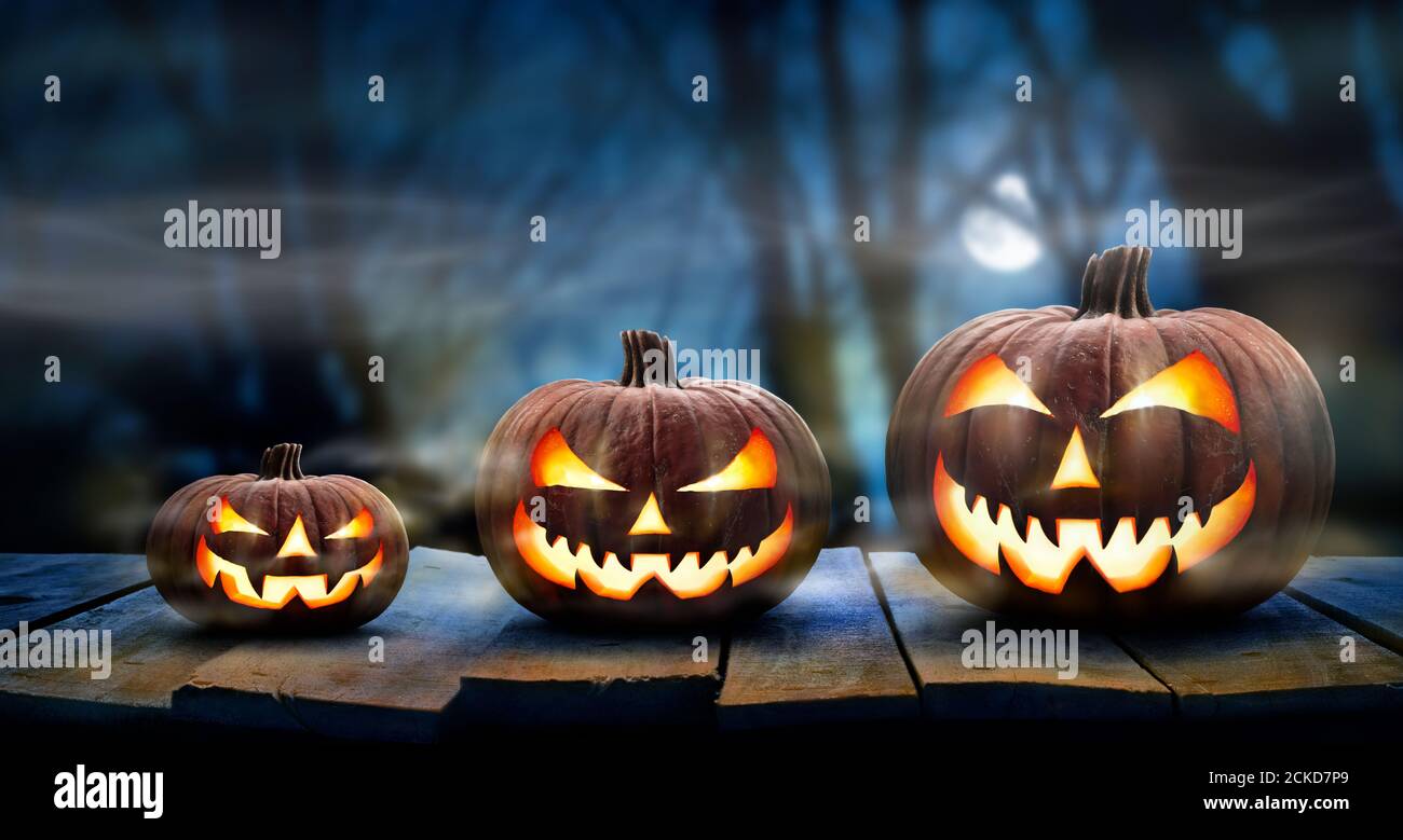 Three spooky halloween pumpkins in a row, Jack O Lantern, with evil face and eyes on a wooden bench, table with a misty night forest background Stock Photo