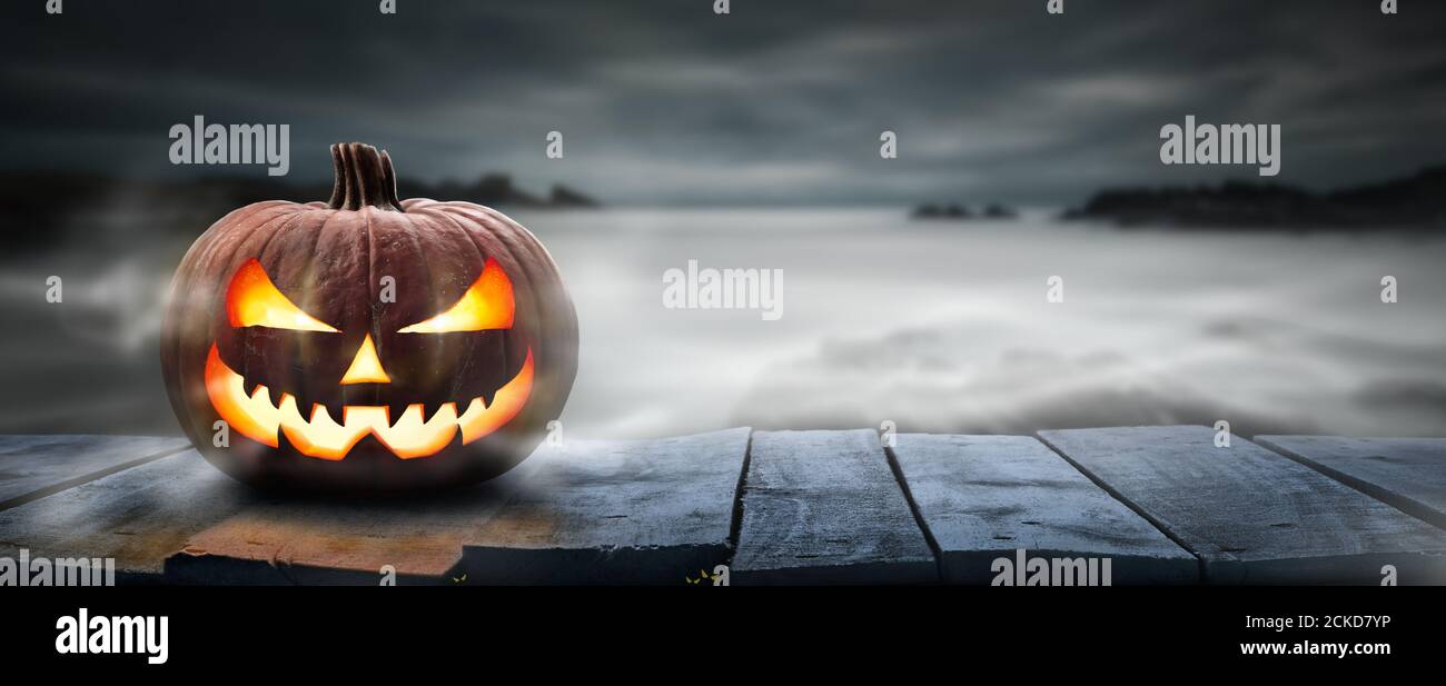 One spooky halloween pumpkin, Jack O Lantern, with an evil face and eyes on a wooden bench, table with a misty gray coastal night background with spac Stock Photo
