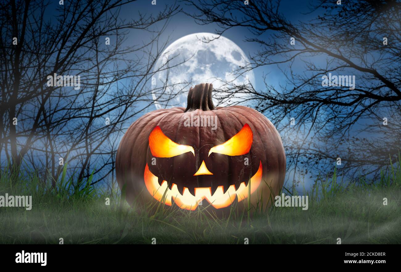 One spooky halloween pumpkin, Jack O Lantern, with an evil face and eyes on the grass with a misty night sky background with a full moon. Stock Photo