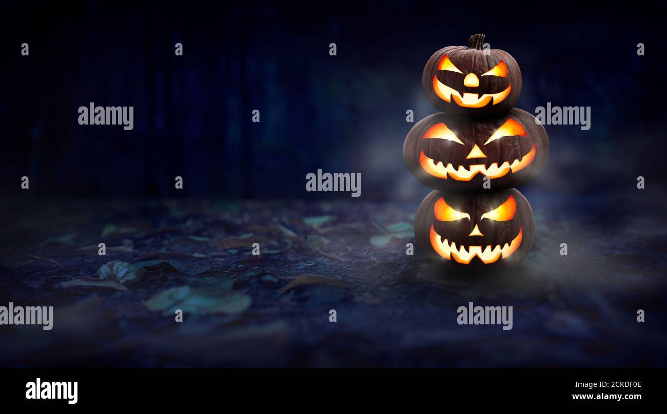 A stack of 3 spooky halloween pumpkin, Jack O Lantern, with an evil face and eyes on the forest floor at night with a dark background. Stock Photo