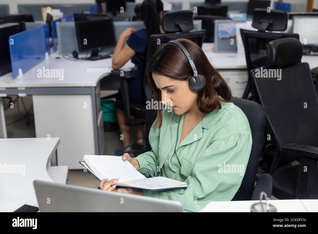 portrait-of-pretty-indian-businesswoman-wearing-headphones-sitting-at-her-workstation-looking-into-her-diary-corporate-environment-call-centre-2CKDFCN.jpg