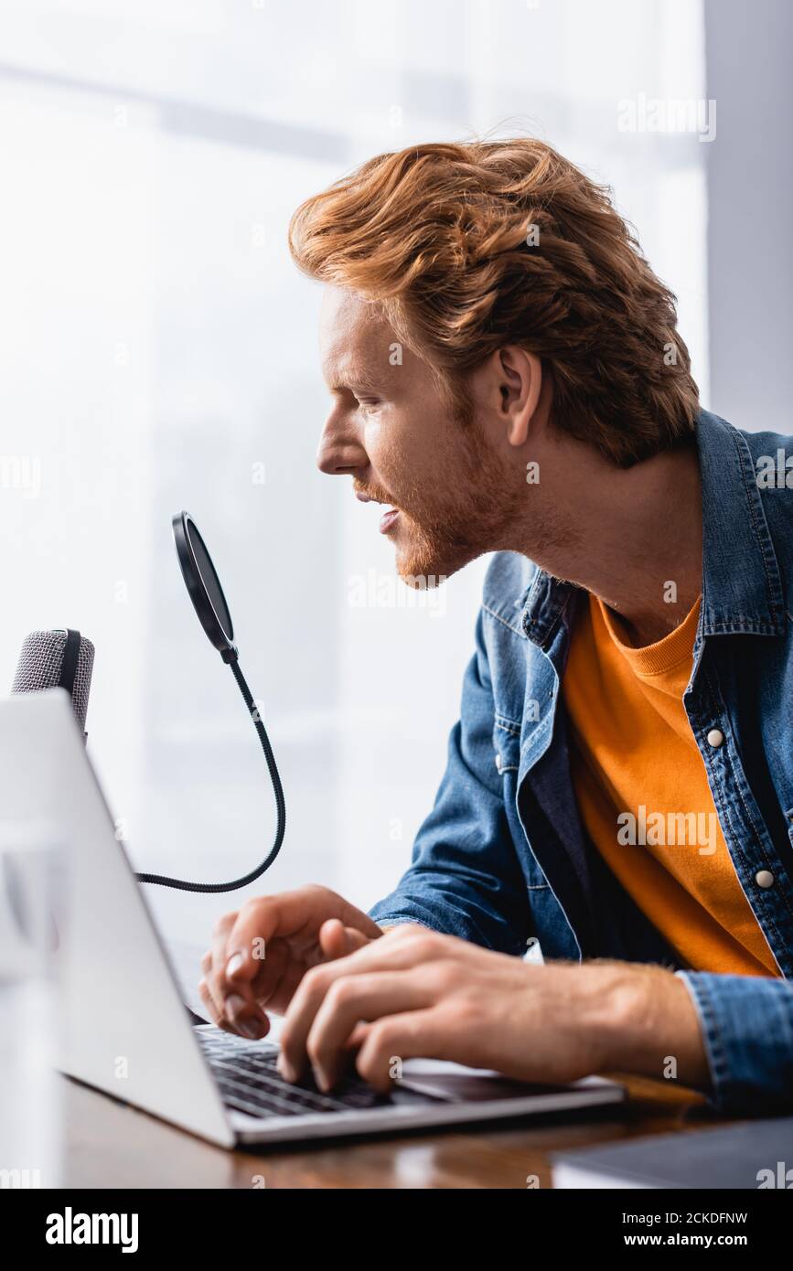 focused, redhead broadcaster speaking in microphone while using laptop Stock Photo