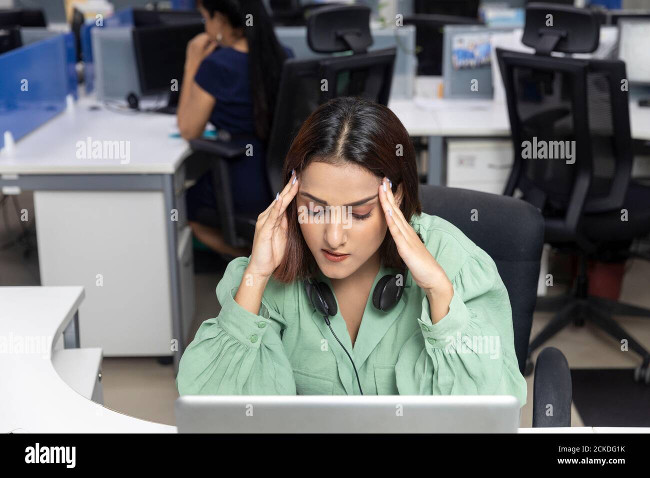 Portrait of tired Indian businesswoman wearing headphones, sitting at ...