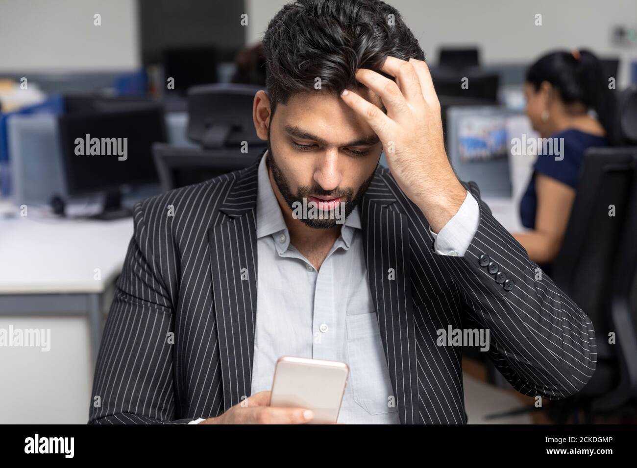 Portrait of young Indian businessman  sitting at his workstation against office background, checking his phone, scratching his head, Stock Photo