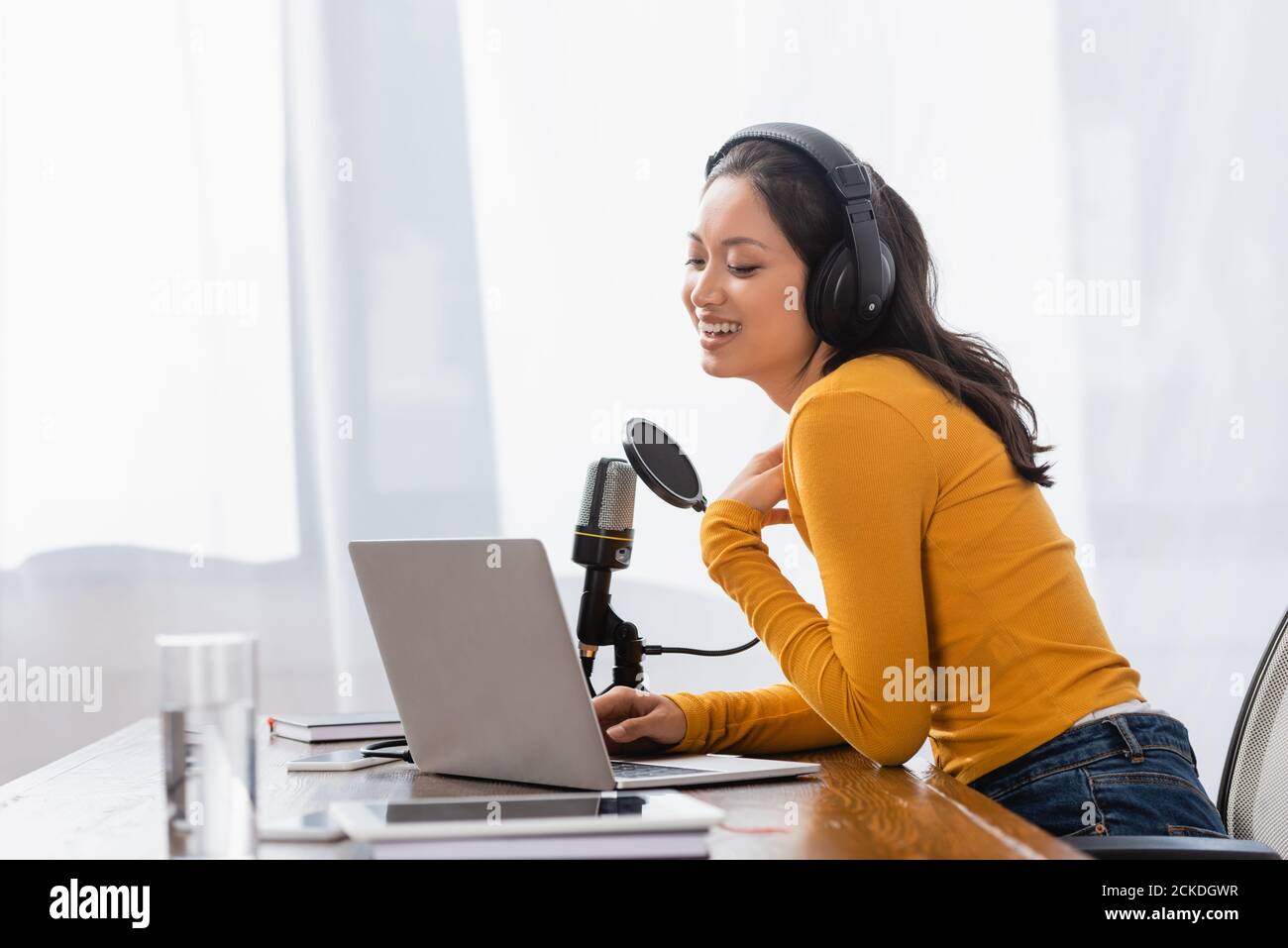 laughing asian radio host in wireless headphones touching chest while working near microphone and laptop Stock Photo