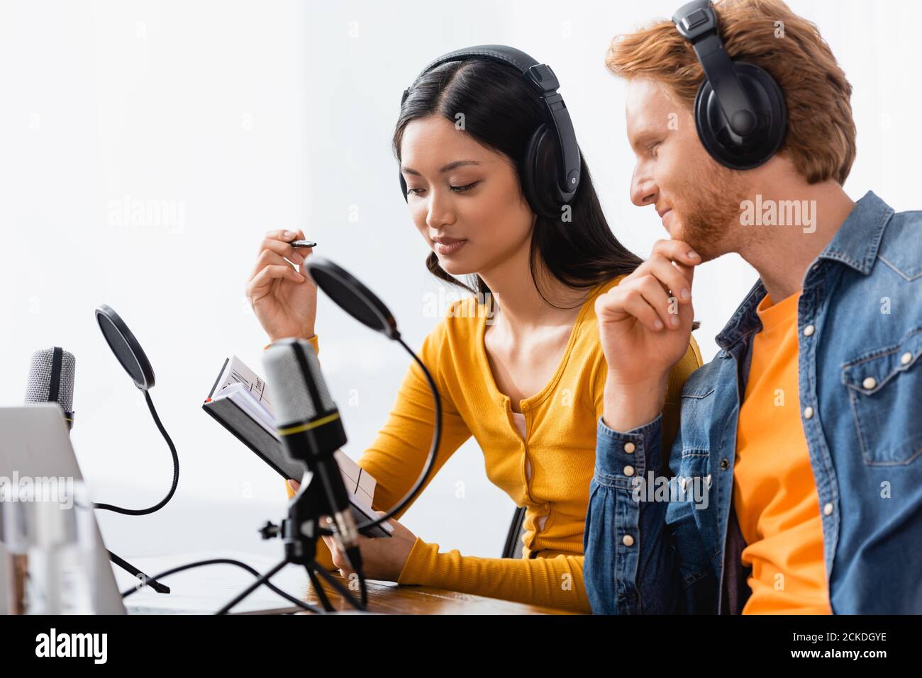 interracial couple of broadcasters in wireless headphones looking at notebook near microphones in studio Stock Photo