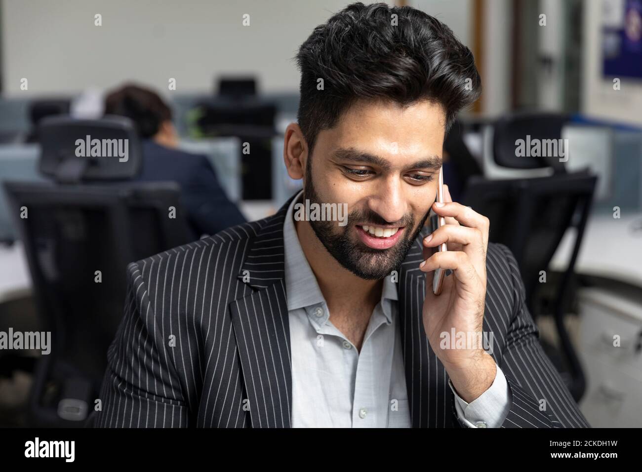Portrait of young Indian businessman speaking on his phone, sitting at his workstation against office background, corporate environment. Stock Photo