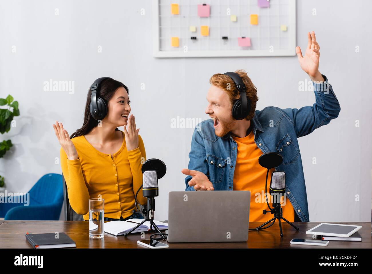 excited multicultural broadcasters in wireless headphones gesturing and looking at each other at workplace Stock Photo
