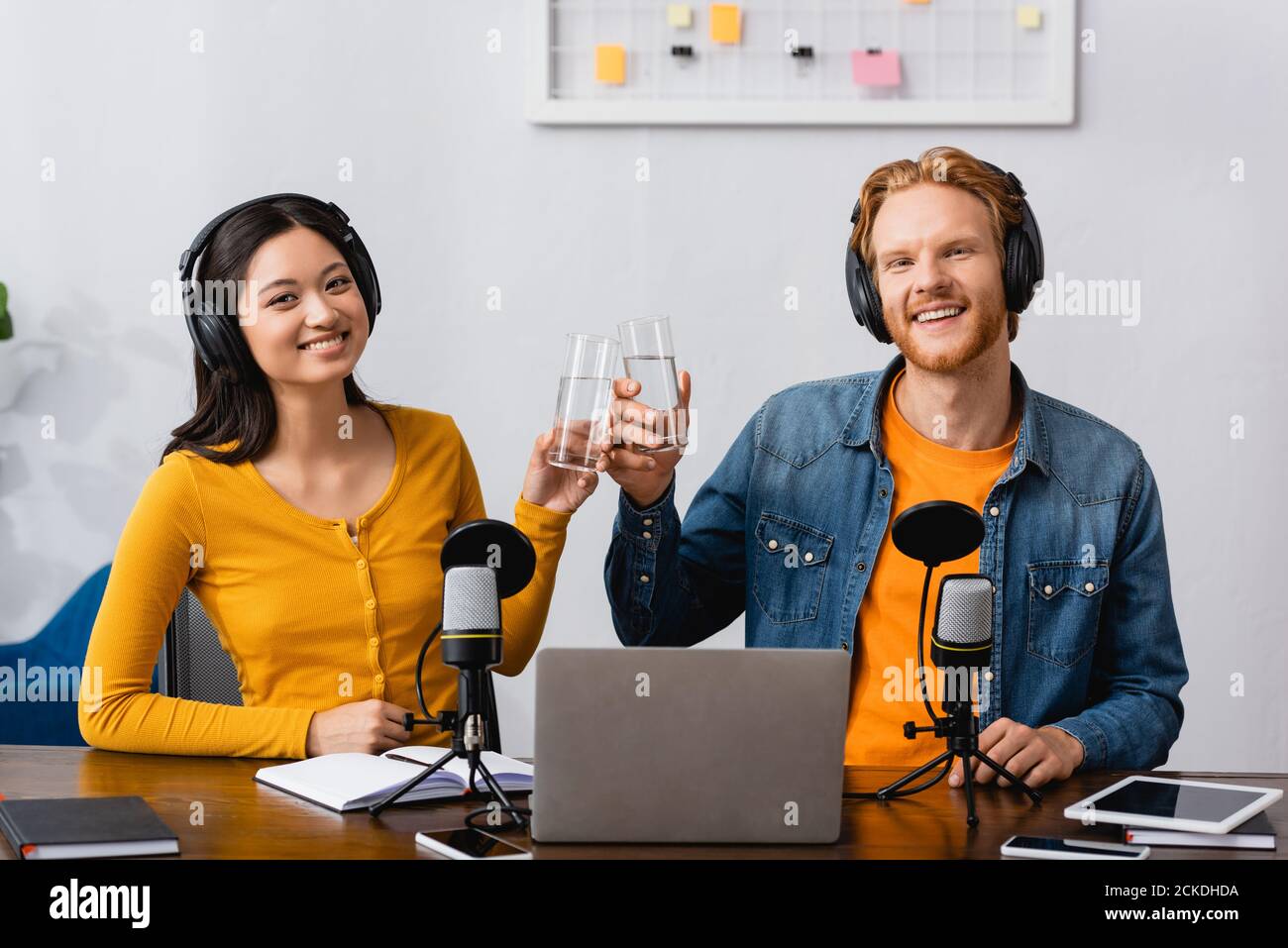 interracial couple of broadcasters in wireless headphones clinking glasses of water while looking at camera Stock Photo