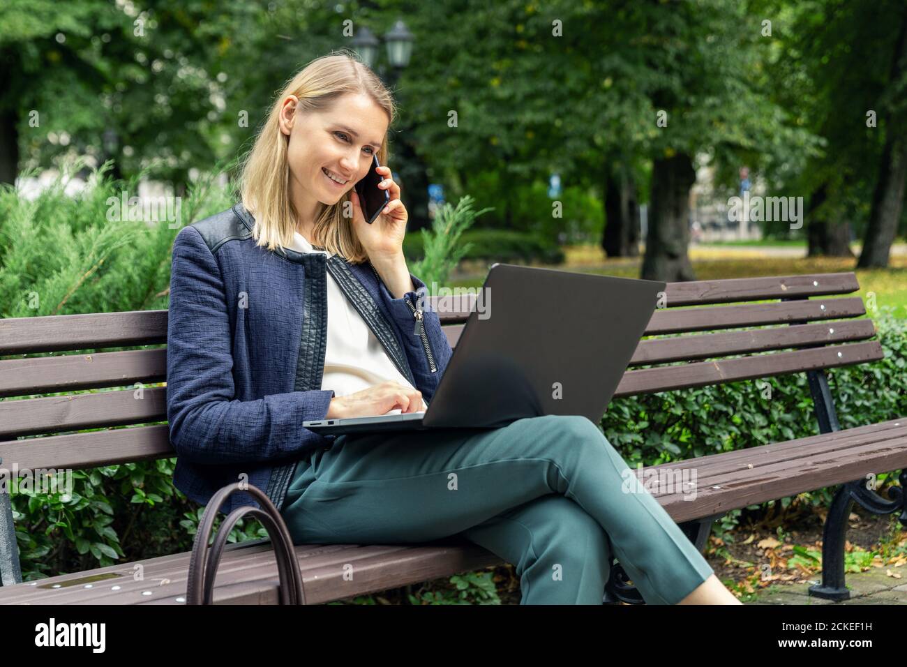 young happy woman sitting on the bench in the city park and talking on the mobile phone while using laptop Stock Photo