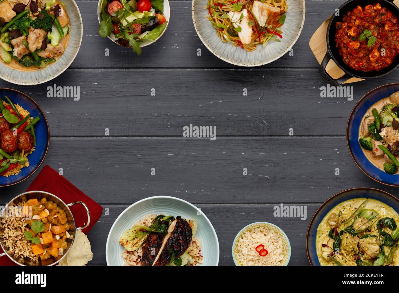 A border of bowls of delicious asian food, curries, chicken chow mein noodles, balti dishes, vegetable curries, on a dark wooden background with space Stock Photo