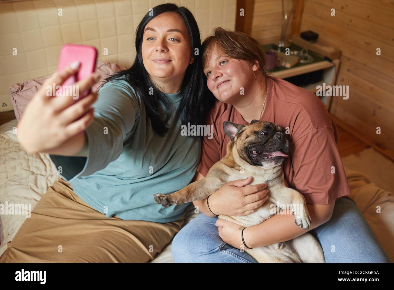 Two overweight women sitting on bed and making selfie portrait on mobile phone with their pet Stock Photo