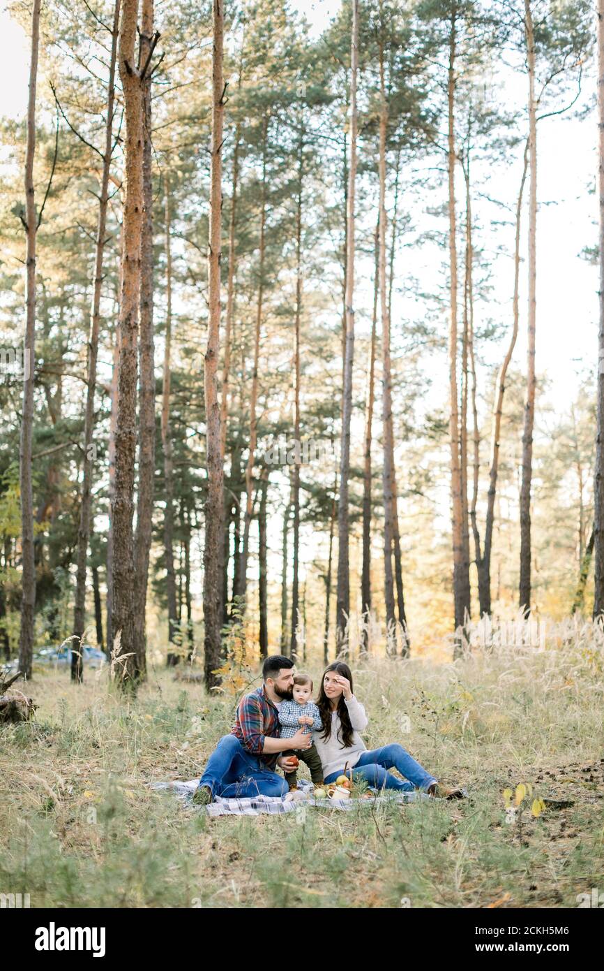 Lifestyle outdoor family portrait of young parents with little kid son, having fun together, while sitting on the blanket in beautiful autumn forest Stock Photo
