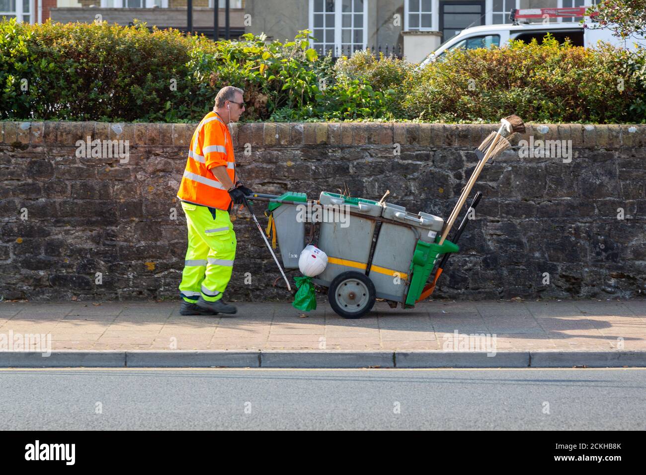 A road sweeper in high visibility clothing pushing his dust cart with brooms in it along the street Stock Photo